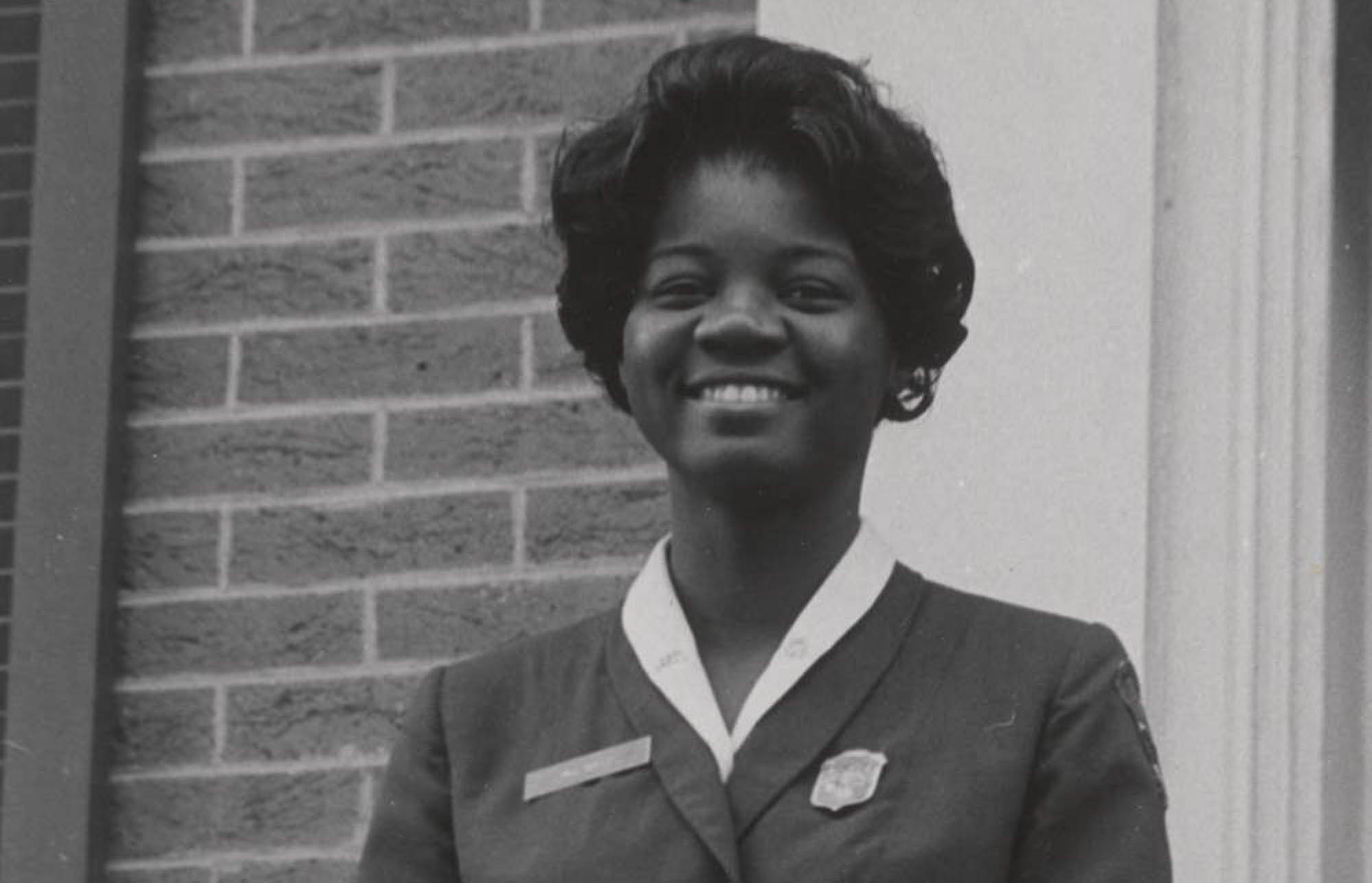 Miss Rolle stands smiling at a railing on top of steps. She wears the NPS women’s uniform skirt, blouse, and jacket with shield-shaped badge.